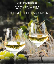 Zwei Weingläser mit weißem Wein stehen auf einem Tisch, umgeben von Trauben. Im Hintergrund sind grüne Blätter sichtbar.