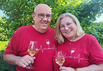 Ein Paar lächelt und hält Weingläser in der Hand. Beide tragen rote T-Shirts mit einem Weinglasmotiv.