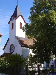 Eine charmante Kirche mit einem schlanken Turm und einer großen Fensterrose. Umgeben von Bäumen und grünem Gestrüpp.