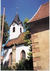 Eine kleine Kirche mit einem hohen Turm und einem charakteristischen Dach. Umgeben von Bäumen und einem alten Gebäude.