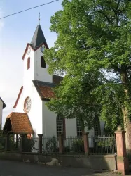Eine alte Kirche mit einem spitzen Turm und großen Fenstern. Im Vordergrund steht ein großer Baum, der Schatten spendet.