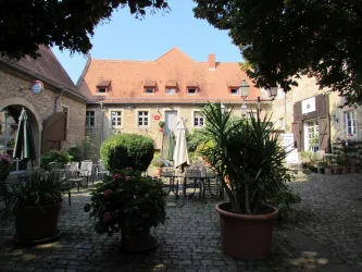 Ein gemütlicher Innenhof mit Pflanzen und Tischen. Im Hintergrund steht ein historisches Gebäude mit rotem Dach.