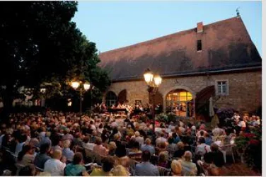Eine große Menschenmenge sitzt vor einer historischen Halle bei Dämmerung. Im Hintergrund findet ein Konzert statt, das von Laternen beleuchtet wird.