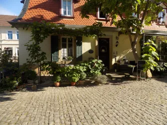 Ein charmantes Haus mit einem roten Dach und grünen Fensterläden. Der Eingangsbereich ist von Pflanzen und einem gepflasterten Weg umgeben.