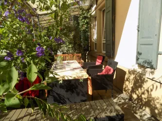 Ein gemütlicher Außenbereich mit einem Tisch und Stühlen, umgeben von Pflanzen. Die Sonne scheint und sorgt für eine einladende Atmosphäre.
