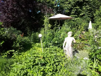 Ein üppiger Garten mit vielen Pflanzen und Sträuchern. In der Mitte steht eine Statue und ein Sonnenschirm bietet Schatten.