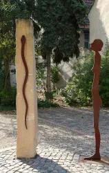 Eine künstlerische Installation mit einer schlanken Figur aus Metall und einem hohen, vertikalen Stein. Im Hintergrund sind Bäume und eine grüne Umgebung zu sehen.