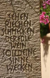 Eine steinerne Wand mit eingravierten Worten. Im Hintergrund sind blühende Pflanzen sichtbar.