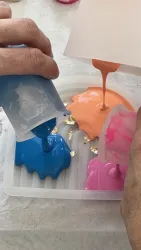 Zwei Hände gießen bunte Farbe in eine Form. Die Farben sind blau, orange und pink und laufen ineinander.