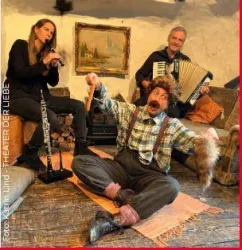 Eine humorvolle Szene im Theater mit drei Darstellern. Ein Mann in Kostüm sitzt auf dem Boden, während zwei Musiker im Hintergrund spielen.