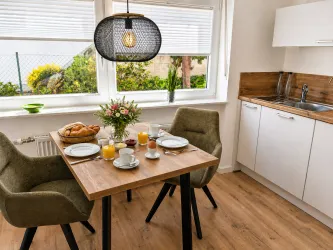 Ein einladender Frühstückstisch mit frischen Brötchen, Blumen und Getränken. Helle, moderne Küche mit großem Fenster und gemütlichen Stühlen.