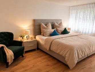 Ein stilvolles Schlafzimmer mit einem großen Bett und mehreren Kissen. Eine gemütliche Leseecke mit einem grünen Sessel ergänzt den Raum.
