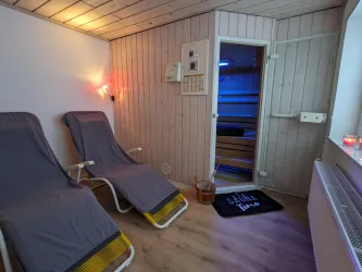 Ein entspannender Raum mit zwei Liegen und einer Sauna. Sanftes Licht schafft eine gemütliche Atmosphäre.