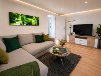 Ein modernes Wohnzimmer mit einer hellen Couch und grünen Kissen. Ein Fernseher und ein Tisch mit einer kleinen Pflanze bringen eine gemütliche Atmosphäre.