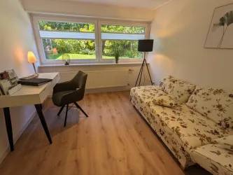 Ein einladendes Zimmer mit einem blühenden Sofa und einem Schreibtisch. Großen Fenstern bieten einen Blick auf den Garten.