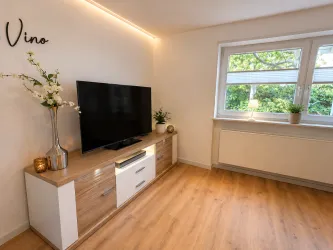 Ein modernes Wohnzimmer mit einem Fernseher auf einer Holzkommode. Die Einrichtung ist hell und freundlich mit Pflanzen und viel natürlichem Licht.