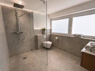 Ein modernes Badezimmer mit einer Glasdusche und eleganten Fliesen. Es gibt auch frische Handtücher und große Fenster, die für Tageslicht sorgen.