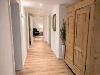Ein heller Flur mit Holzfußboden und weißen Wänden. Am Ende des Flurs steht ein bequemer Stuhl neben einem Schrank aus Holz.