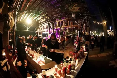 Ein festlicher Weihnachtsmarkt mit bunten Lichtern und vielen Menschen. Stände bieten Kerzen und Dekorationen an.