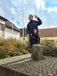 Ein Mann steht auf einem Platz und hält eine Marionette in der Hand. Im Hintergrund sind Blumen und Häuser zu sehen.