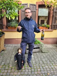 Ein Mann steht auf einem Pflasterboden und hält eine Flasche Sekt in der linken Hand. In der rechten Hand führt er eine Marionette mit einem kleinen Mann in schwarzer Kleidung.
