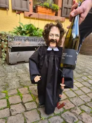 Eine kleine Puppe in einem Richtergewand hält eine Flasche Wein. Im Hintergrund sind Pflanzen und ein Holzkasten sichtbar.