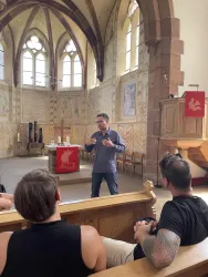 Ein Mann spricht in einer Kirche, während Zuhörer auf einer Holzbank sitzen. Der Raum ist hell erleuchtet und zeigt ansprechende Wandmalereien.
