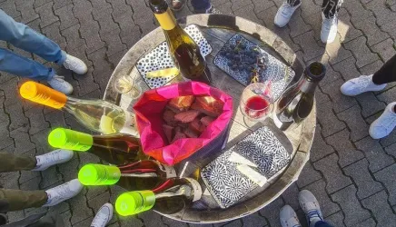 Ein Tisch mit verschiedenen Weinen, Snacks und Gläsern. Um den Tisch stehen mehrere Personen in sportlichen Schuhen.