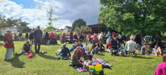 Eine große Gruppe von Menschen versammelt sich auf einer Wiese. Es gibt viele Picknickdecken und Bäume im Hintergrund.