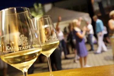 <p>Zwei Weingläser mit Aufschrift Winzergenossenschaft Kallstadt und  Gästen im Hintergrund</p>
