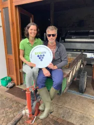 Ein Paar sitzt auf einem Wagen vor einem Weingut und hält ein Zeichen von ECOVIN. Im Hintergrund sind Geräte zur Weinproduktion sichtbar.