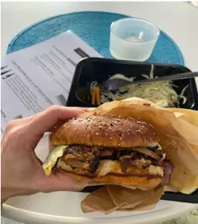 Ein saftiger Burger mit Fleisch, Salat und Sesambrötchen wird in der Hand gehalten. Im Hintergrund sind ein Glas Wasser und eine Schale mit Beilage zu sehen.