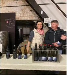 Zwei Personen stehen hinter einem Tisch mit Weinflaschen. Im Hintergrund sind Weinfässer und eine Tafel mit Informationen zu sehen.