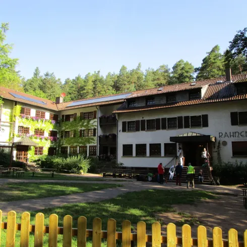 Ein zweigeschossiges Gebäude mit bunten Fassaden umgeben von Bäumen. Vor dem Gebäude sind Bänke und ein gepflegter Rasen zu sehen.