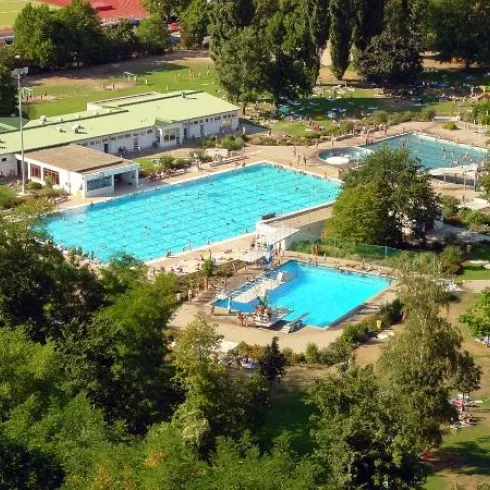 Ein Schwimmbad mit mehreren Becken und viel grünem Gartenbereich. Das Gelände ist von Bäumen umgeben und es sind viele Besucher zu sehen.