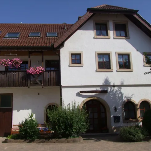 Ein schönes Haus mit einem Balkon, auf dem Blumentöpfe stehen. Die Fassade ist weiß und der Dachbereich braun, umgeben von grünen Pflanzen.