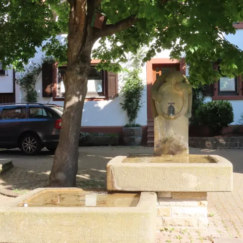 <p>Bobenheimer Winzerbrunnen mit zwei Wasserbecken aus Sandstein und einer Figur obenauf. Auf der linken Seite beschattet ein Baum den Brunnen, im Hintergrund ist ein Haus  und ein Auto zu sehen.