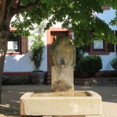 <p>Bobenheimer Winzerbrunnen Vorderansicht, beschattet von einem Baum. Sandsteinbrunnen mit Figur und einem Haus im Hintergrund. </p>