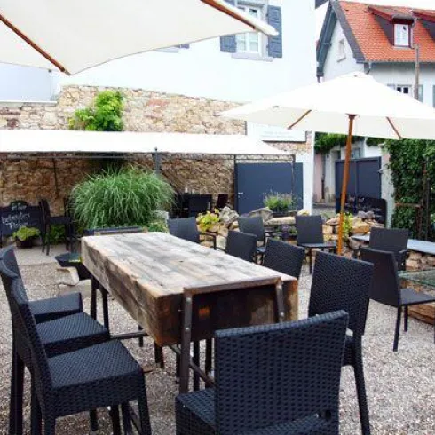 Ein gemütlicher Außenbereich mit einem großen Tisch und schwarzen Stühlen. Sonnenschirme bieten Schatten, umgeben von grünen Pflanzen und einer rustikalen Steinmauer.