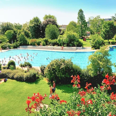 Ein schöner Pool umgeben von Grünanlagen und Blumen. Menschen genießen das Wasser und entspannen in der Sonne.