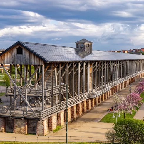 Ein historisches Holzgebäude mit einer offenen Struktur steht in einer malerischen Umgebung. Umgeben von blühenden Bäumen und einer weitläufigen Grünfläche.