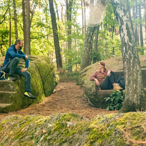 Zwei Personen sitzen auf alten Steinen im Wald. Die Umgebung ist grün und sonnig, umgeben von Bäumen.