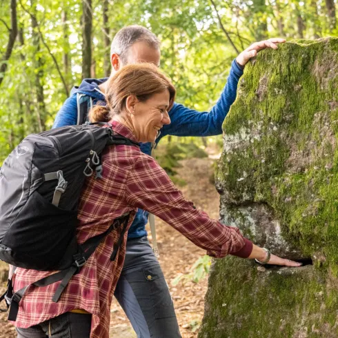 Ein Paar erkundet einen Waldweg und betrachtet einen moosbedeckten Felsen. Die Atmosphäre ist naturverbunden und abenteuerlich.