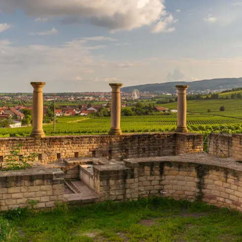 Eine Aussicht auf Weinberge mit alten Säulen im Vordergrund. Der Himmel ist teilweise bewölkt und die Landschaft wirkt friedlich.