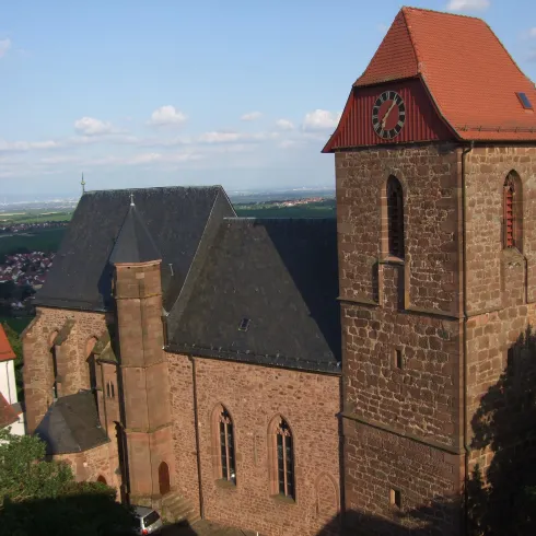 Eine alte Kirche mit einem rot gedeckten Dach und einem Glockenturm. Die Fassade ist aus Stein und umgeben von einer grünen Landschaft.