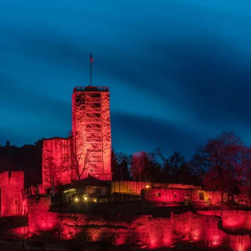Eine beleuchtete Burg bei Nacht mit rotem Licht, umgeben von Bäumen. Der Himmel ist blau und bewölkt.