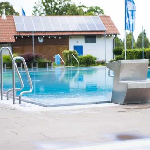 Ein klarer Pool steht vor einem Gebäude mit Solarzellen auf dem Dach. Die Umgebung ist gepflegt und mit grünen Pflanzen ausgestattet.