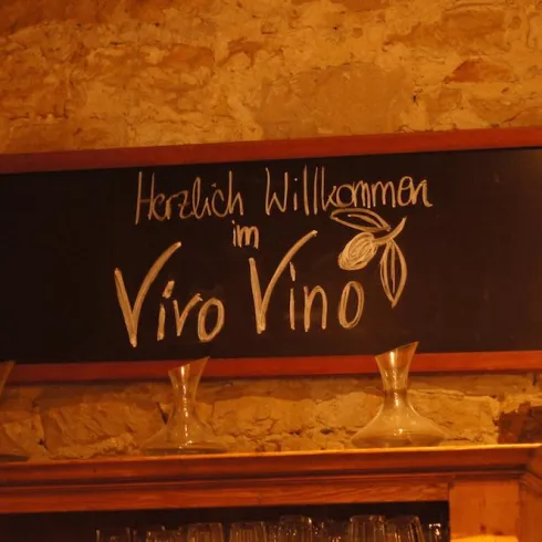 Ein Willkommensschild für das Vino Vino. Die Wand ist aus Stein, und es stehen einige Gläser bereit.