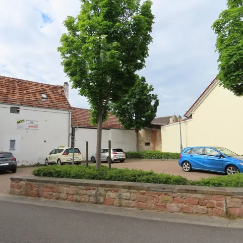 Eine kleine Mauer die den Parkplatz von der Straße abtrennt. Daneben sind einzelne Bäume als Deko.