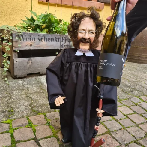 Eine kleine Puppe in einem Richtergewand hält eine Flasche Wein. Im Hintergrund sind Pflanzen und ein Holzkasten sichtbar.